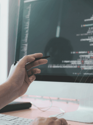 Développeur travaillant sur un ordinateur double écran affichant du code source, avec une main tenant un stylet pointant vers l'écran principal. Atmosphère de bureau technique et logo "dcod" visible en bas à droite.