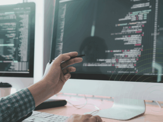 Développeur travaillant sur un ordinateur double écran affichant du code source, avec une main tenant un stylet pointant vers l'écran principal. Atmosphère de bureau technique et logo "dcod" visible en bas à droite.