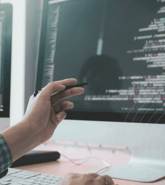 Développeur travaillant sur un ordinateur double écran affichant du code source, avec une main tenant un stylet pointant vers l'écran principal. Atmosphère de bureau technique et logo "dcod" visible en bas à droite.