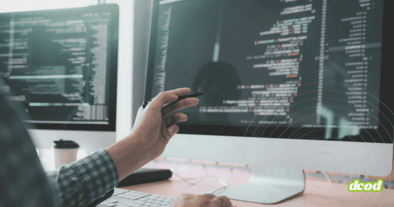 Développeur travaillant sur un ordinateur double écran affichant du code source, avec une main tenant un stylet pointant vers l'écran principal. Atmosphère de bureau technique et logo "dcod" visible en bas à droite.