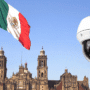 Grand drapeau mexicain flottant devant la cathédrale métropolitaine de Mexico, avec en premier plan une caméra de vidéosurveillance moderne illustrant l’essor des technologies de sécurité dans l’espace public.