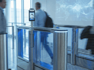 Des passagers flous franchissent des portiques de sécurité automatisés équipés d'écrans de reconnaissance faciale dans un aéroport.