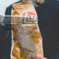 Montage photographique représentant un paquet de chips Lay’s saveur poulet rôti tenu par une personne en costume, avec une superposition d’image d’un pistolet semi-automatique, créant un contraste visuel fort entre aliment et arme à feu.