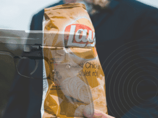 Montage photographique représentant un paquet de chips Lay’s saveur poulet rôti tenu par une personne en costume, avec une superposition d’image d’un pistolet semi-automatique, créant un contraste visuel fort entre aliment et arme à feu.