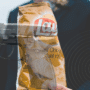 Un système AI confond un paquet de chips avec une arme dans un lycée 1 Montage photographique représentant un paquet de chips Lay’s saveur poulet rôti tenu par une personne en costume, avec une superposition d’image d’un pistolet semi-automatique, créant un contraste visuel fort entre aliment et arme à feu.