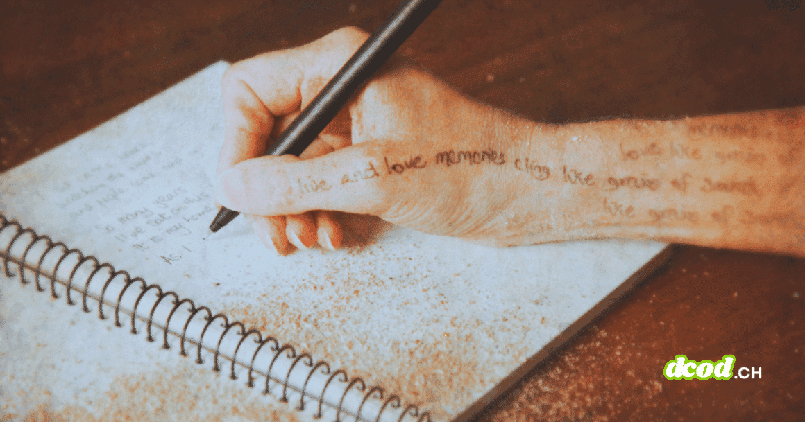 Une main tenant un stylo écrit dans un carnet spiralé posé sur une table, avec du sable répandu sur la page et sur la peau.