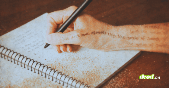 Une main tenant un stylo écrit dans un carnet spiralé posé sur une table, avec du sable répandu sur la page et sur la peau.