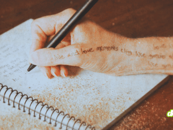 Une main tenant un stylo écrit dans un carnet spiralé posé sur une table, avec du sable répandu sur la page et sur la peau.