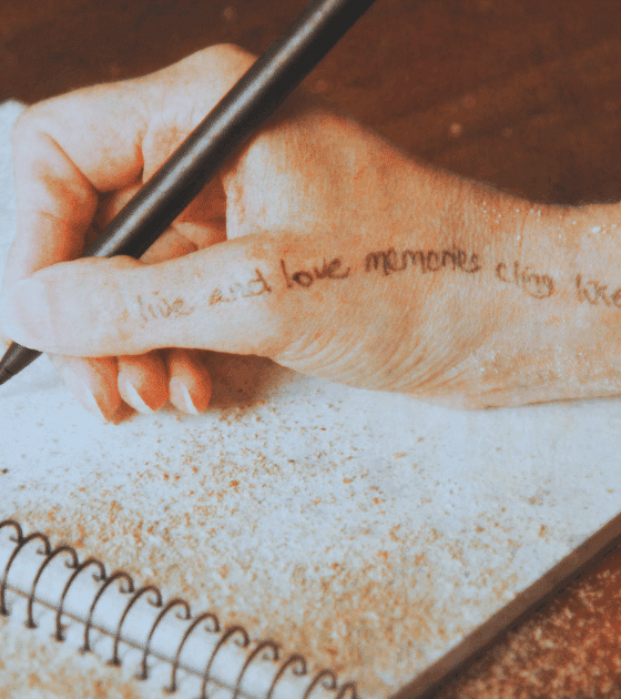 Une main tenant un stylo écrit dans un carnet spiralé posé sur une table, avec du sable répandu sur la page et sur la peau.