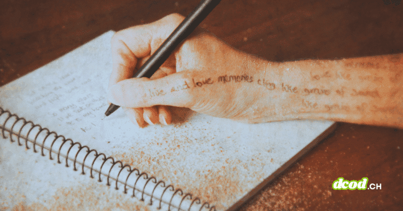 Une main tenant un stylo écrit dans un carnet spiralé posé sur une table, avec du sable répandu sur la page et sur la peau.