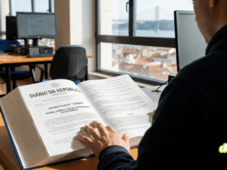 Un homme vu de dos, vêtu d'un sweat-shirt à capuche bleu marine, est assis à un bureau et lit un grand livre ouvert. Le livre est le "DIÁRIO DA REPÚBLICA" (Journal Officiel portugais), et la page de droite présente le titre "DECRETO-LEI N.º 125/2023 - REGIME JURÍDICO DOS INVESTIGADORES DE SEGURANÇA". Sa main est posée sur les pages. La scène se déroule dans un bureau ou une salle informatique avec des ordinateurs de bureau inoccupés. Par une grande fenêtre en arrière-plan, on aperçoit une vue panoramique de Lisbonne et le pont du 25 Avril sous un ciel clair.