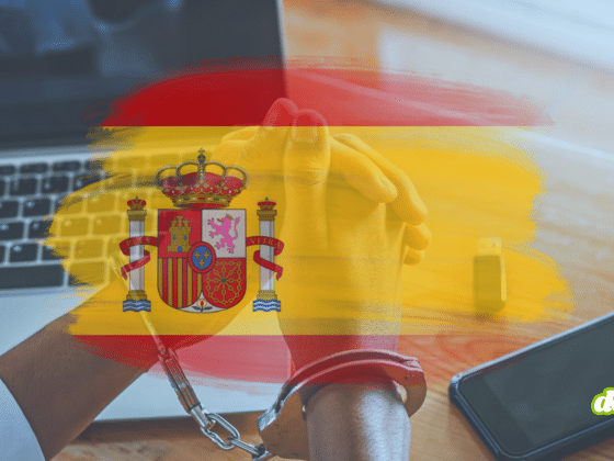 Mains menottées devant un ordinateur portable, avec drapeau espagnol en surimpression et smartphone sur la table.