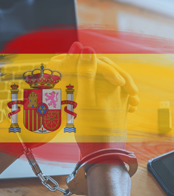Mains menottées devant un ordinateur portable, avec drapeau espagnol en surimpression et smartphone sur la table.