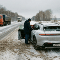 Un homme en manteau foncé et bonnet noir se tient à côté de la portière conducteur ouverte d'une Porsche Taycan grise, garée sur le bas-côté enneigé d'une route de campagne en hiver. La voiture porte une plaque d'immatriculation russe. Un camion orange et un autre véhicule circulent sur la route mouillée et couverte de boue au second plan. La scène est entourée d'une forêt d'arbres dénudés sous un ciel gris.