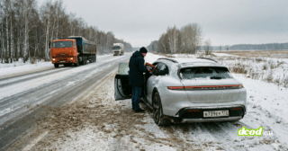 Un homme en manteau foncé et bonnet noir se tient à côté de la portière conducteur ouverte d'une Porsche Taycan grise, garée sur le bas-côté enneigé d'une route de campagne en hiver. La voiture porte une plaque d'immatriculation russe. Un camion orange et un autre véhicule circulent sur la route mouillée et couverte de boue au second plan. La scène est entourée d'une forêt d'arbres dénudés sous un ciel gris.