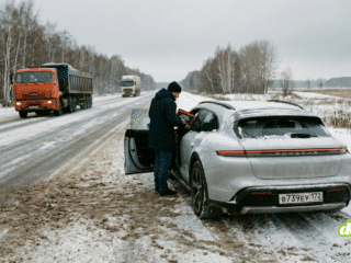 Un homme en manteau foncé et bonnet noir se tient à côté de la portière conducteur ouverte d'une Porsche Taycan grise, garée sur le bas-côté enneigé d'une route de campagne en hiver. La voiture porte une plaque d'immatriculation russe. Un camion orange et un autre véhicule circulent sur la route mouillée et couverte de boue au second plan. La scène est entourée d'une forêt d'arbres dénudés sous un ciel gris.