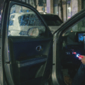Image nocturne montrant une personne à l'intérieur d'une Hyundai Ioniq 5, connectant un appareil à un ordinateur portable affichant du code. Une Kia EV6 est garée juste devant, et une voiture de police est visible en arrière-plan sur une rue mouillée. Cette scène illustre le problème de la faille de sécurité USB affectant certains véhicules Hyundai et Kia.