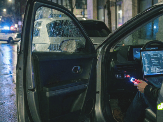 Image nocturne montrant une personne à l'intérieur d'une Hyundai Ioniq 5, connectant un appareil à un ordinateur portable affichant du code. Une Kia EV6 est garée juste devant, et une voiture de police est visible en arrière-plan sur une rue mouillée. Cette scène illustre le problème de la faille de sécurité USB affectant certains véhicules Hyundai et Kia.