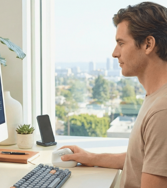 Un homme dans un bureau moderne et lumineux utilise une souris ergonomique verticale Logitech Lift blanche de la main droite tout en regardant des graphiques financiers et des tableaux de bord sur un écran iMac. Son bureau comprend également un clavier mécanique, une tasse de café, une plante en pot, un carnet et un support de téléphone. Une grande baie vitrée offre une vue panoramique sur la ville.