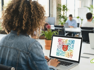 Une femme de dos à un bureau tape sur un ordinateur portable. L'écran affiche un grand QR code multicolore avec un emoji diable rouge au centre, illustrant un lien malveillant.