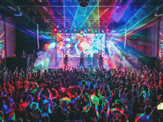 Foule dansant lors d'un concert animé, la scène et les murs étant éclairés par des projections lumineuses psychédéliques et des faisceaux laser de toutes les couleurs.