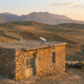 Une petite maison rurale en pierre et en terre dans un paysage vallonné iranien au coucher du soleil, équipée d'une antenne parabolique Starlink sur son toit plat, avec un berger et son troupeau à proximité.