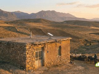Une petite maison rurale en pierre et en terre dans un paysage vallonné iranien au coucher du soleil, équipée d'une antenne parabolique Starlink sur son toit plat, avec un berger et son troupeau à proximité.