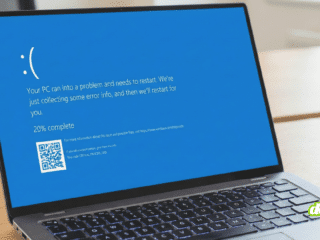 Gros plan sur un ordinateur portable argenté ouvert, posé sur un bureau en bois. L'écran affiche le message d'erreur "écran bleu" de Windows 10/11 avec un grand émoticône triste ":(", le texte "Votre PC a rencontré un problème et doit redémarrer", une progression indiquant "20% effectués", un QR code pour plus d'informations et le code d'arrêt spécifique "CRITICAL_PROCESS_DIED". Le clavier rétroéclairé est visible et l'arrière-plan du bureau est flou.