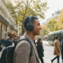 Un homme noir souriant porte des écouteurs Sony WH-1000XM4 et un sac à dos, marchant dans une rue parisienne animée avec un tramway.