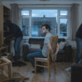 Une photographie en format paysage 16/9 montrant un homme ligoté et bâillonné sur une chaise au centre d'un salon en désordre. Deux individus encagoulés fouillent les meubles de la pièce. Le décor est sombre et l'éclairage faible, suggérant le crépuscule.