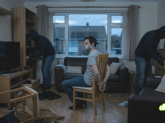 Une photographie en format paysage 16/9 montrant un homme ligoté et bâillonné sur une chaise au centre d'un salon en désordre. Deux individus encagoulés fouillent les meubles de la pièce. Le décor est sombre et l'éclairage faible, suggérant le crépuscule.