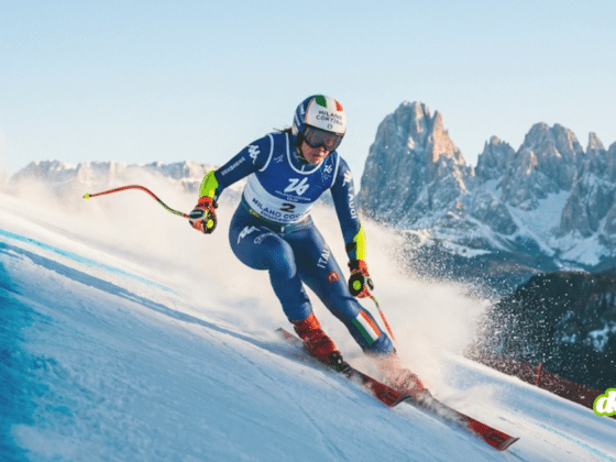 Photographie d'une skieuse alpine italienne en pleine action lors d'une descente de slalom géant, portant le dossard de Milan Cortina 2026, avec les montagnes des Dolomites en arrière-plan.