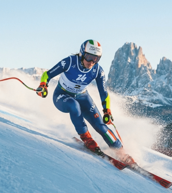 Photographie d'une skieuse alpine italienne en pleine action lors d'une descente de slalom géant, portant le dossard de Milan Cortina 2026, avec les montagnes des Dolomites en arrière-plan.