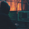 Photographie d'illustration pour la veille cyberattaque : une silhouette portant un sweat à capuche noir est assise de dos devant plusieurs écrans d'ordinateur affichant du code vert complexe et des données. L'environnement est une salle serveur sombre, éclairée par les lueurs bleues des écrans et des lumières oranges en arrière-plan, évoquant un hacker ou un analyste en action.