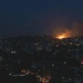 Une photographie nocturne d'un panorama urbain où la plupart des bâtiments sont plongés dans l'obscurité, avec seulement quelques lumières éparses visibles aux fenêtres et dans les rues. Au loin, sur une colline, un important incendie est en cours, dégageant une vive lueur orange et un panache de fumée qui s'élève dans le ciel sombre. Les silhouettes des immeubles se découpent contre le ciel nocturne et la lumière du feu.