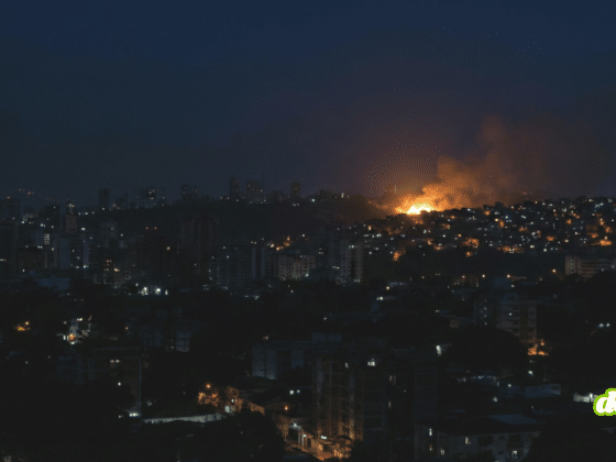 Une photographie nocturne d'un panorama urbain où la plupart des bâtiments sont plongés dans l'obscurité, avec seulement quelques lumières éparses visibles aux fenêtres et dans les rues. Au loin, sur une colline, un important incendie est en cours, dégageant une vive lueur orange et un panache de fumée qui s'élève dans le ciel sombre. Les silhouettes des immeubles se découpent contre le ciel nocturne et la lumière du feu.