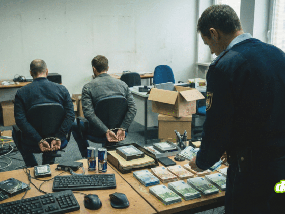 Un policier compte des liasses de billets en euros sur un bureau devant deux hommes menottés dans un bureau en désordre.