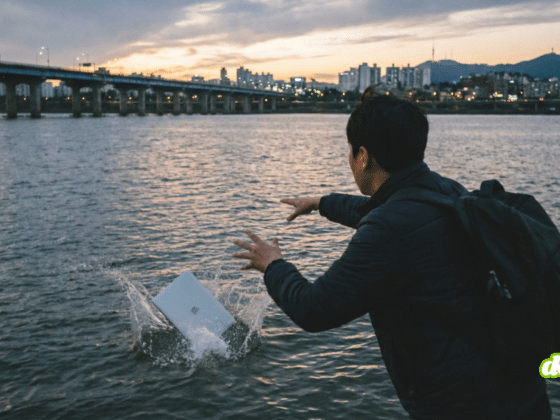 Une photographie au crépuscule montre une personne vue de dos, portant un blouson sombre et un sac à dos, qui vient de jeter un ordinateur portable argenté dans une large rivière. Le MacBook fait éclabousser l'eau au premier plan. En arrière-plan, un pont illuminé traverse le fleuve, et la silhouette de la ville de Séoul, avec la N Seoul Tower visible, se découpe contre un ciel nuageux aux teintes oranges et bleues. L'atmosphère est sombre et dramatique.