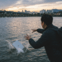 Une photographie au crépuscule montre une personne vue de dos, portant un blouson sombre et un sac à dos, qui vient de jeter un ordinateur portable argenté dans une large rivière. Le MacBook fait éclabousser l'eau au premier plan. En arrière-plan, un pont illuminé traverse le fleuve, et la silhouette de la ville de Séoul, avec la N Seoul Tower visible, se découpe contre un ciel nuageux aux teintes oranges et bleues. L'atmosphère est sombre et dramatique.