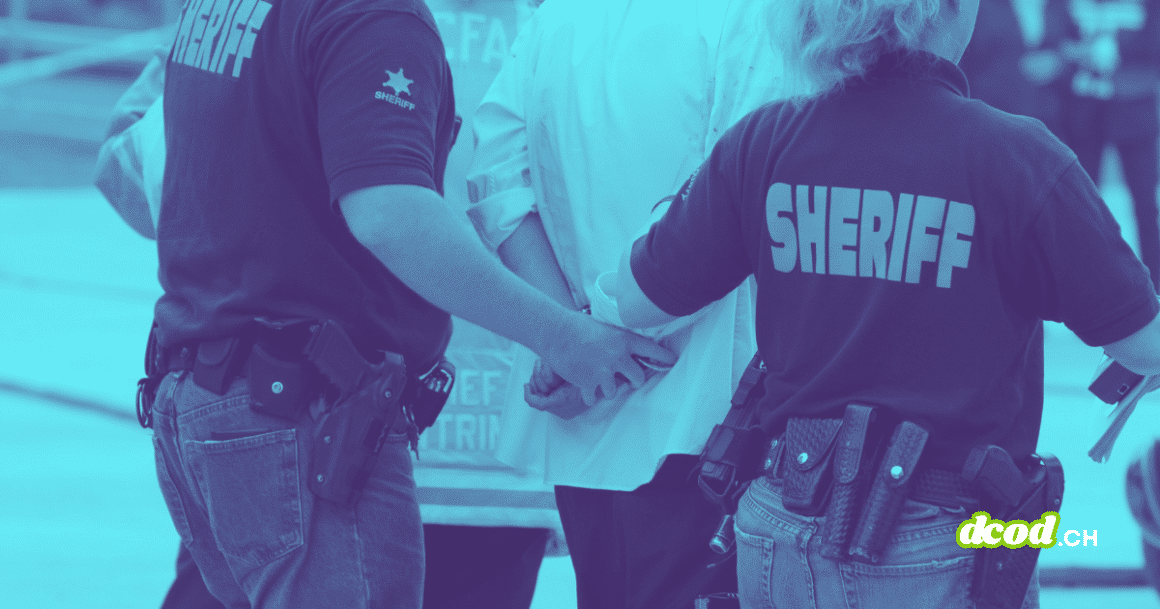 Photographie teintée en bleu montrant deux agents du bureau du shérif, de dos, en train de menotter une personne. On aperçoit leurs uniformes marqués "SHERIFF" et leurs ceinturons d'équipement.