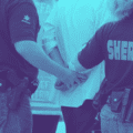 Photographie teintée en bleu montrant deux agents du bureau du shérif, de dos, en train de menotter une personne. On aperçoit leurs uniformes marqués "SHERIFF" et leurs ceinturons d'équipement.