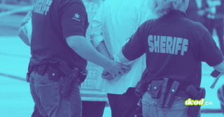 Photographie teintée en bleu montrant deux agents du bureau du shérif, de dos, en train de menotter une personne. On aperçoit leurs uniformes marqués "SHERIFF" et leurs ceinturons d'équipement.