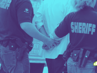 Photographie teintée en bleu montrant deux agents du bureau du shérif, de dos, en train de menotter une personne. On aperçoit leurs uniformes marqués "SHERIFF" et leurs ceinturons d'équipement.