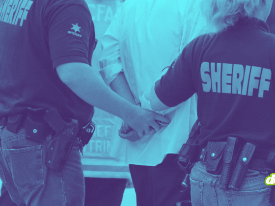 Photographie teintée en bleu montrant deux agents du bureau du shérif, de dos, en train de menotter une personne. On aperçoit leurs uniformes marqués "SHERIFF" et leurs ceinturons d'équipement.