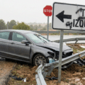 Une voiture autonome grise sans marque identifiable accidentée contre une glissière de sécurité tordue et arrachée sur le bord d'une route mouillée. Au premier plan, un panneau de signalisation blanc pirate affiche l'instruction contradictoire en spanglish « TURN IZQUIERDO » avec une flèche vers la gauche, tandis qu'un panneau STOP réglementaire est visible en arrière-plan, illustrant une erreur de trajectoire causée par une injection de prompt physique.