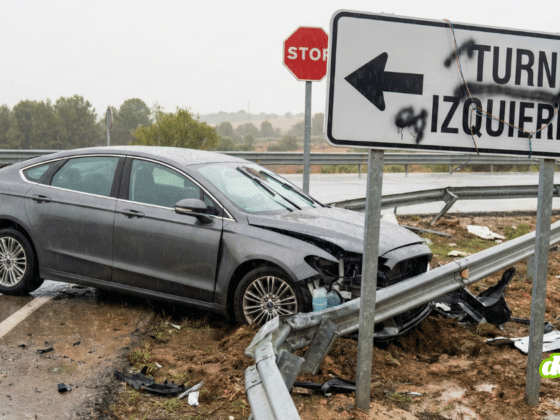 Une voiture autonome grise sans marque identifiable accidentée contre une glissière de sécurité tordue et arrachée sur le bord d'une route mouillée. Au premier plan, un panneau de signalisation blanc pirate affiche l'instruction contradictoire en spanglish « TURN IZQUIERDO » avec une flèche vers la gauche, tandis qu'un panneau STOP réglementaire est visible en arrière-plan, illustrant une erreur de trajectoire causée par une injection de prompt physique.