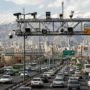 Une vue panoramique en hauteur de l'autoroute Modares à Téhéran, bondée de voitures. Un grand portique métallique traverse la route, supportant de nombreuses caméras de surveillance et radars de vitesse, avec la ville dense et des montagnes enneigées en arrière-plan.