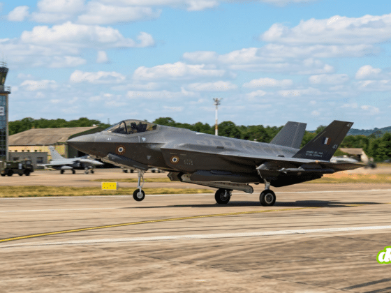 Photographie d'un chasseur furtif F-35 au décollage sur la piste d'une base aérienne avec une tour de contrôle en arrière-plan sous un ciel nuageux.