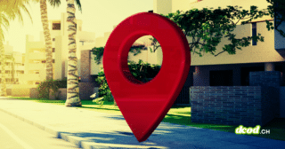 Une grande épingle de géolocalisation rouge posée sur un trottoir devant des bâtiments modernes et des palmiers, illustrant le traçage géographique et la surveillance.