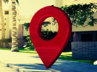 Une grande épingle de géolocalisation rouge posée sur un trottoir devant des bâtiments modernes et des palmiers, illustrant le traçage géographique et la surveillance.
